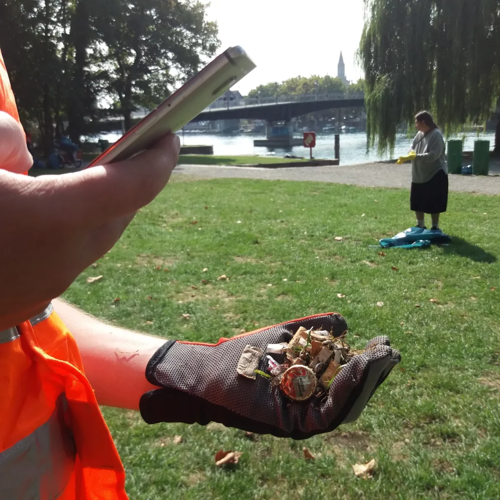 Menschliche Hand mit Schutzhandschuhen hält gesammelten Müll in der Hand, die andere Hand macht mit Smartphone ein Foto davon. Im Hintergrund die Kulisse von Konstanz am Bodensee.