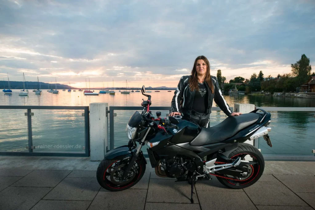 Frau steht hinter ihrem Motorrad vor der Kulisse der untergehenden Sonne am Wasser des Bodensees.