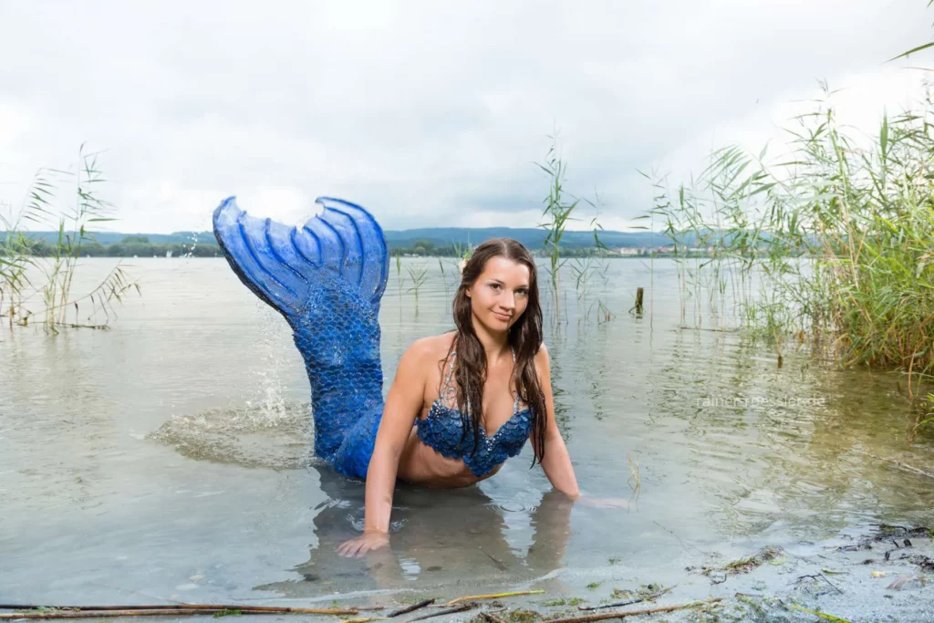 Frau mit blauen Meerjungfrau Kostum im Wasser vom Bodensee. Dichte Wolken am Himmel.