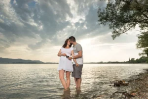 bodensee radolfzell mettnau pärchen frau mann menschen wasser babybauch draussen schwanger sommer fotoshooting 2017 2 Menschen stehen im Wasser und halten ein Ultraschall Foto vor dem Babybauch. Beide Menschen küssen sich vor der Kulisse der untergehenden Sonne im Wasser vom Bodensee. Im Hintergrund ist der Kirchspitze vom Münster in Radolfzell zu sehen.