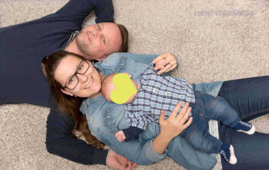 Eltern und Neugeborenes liegen auf dem Boden. Das Gesicht vom Kind ist digital unkenntlich gemacht mit einem Herz.