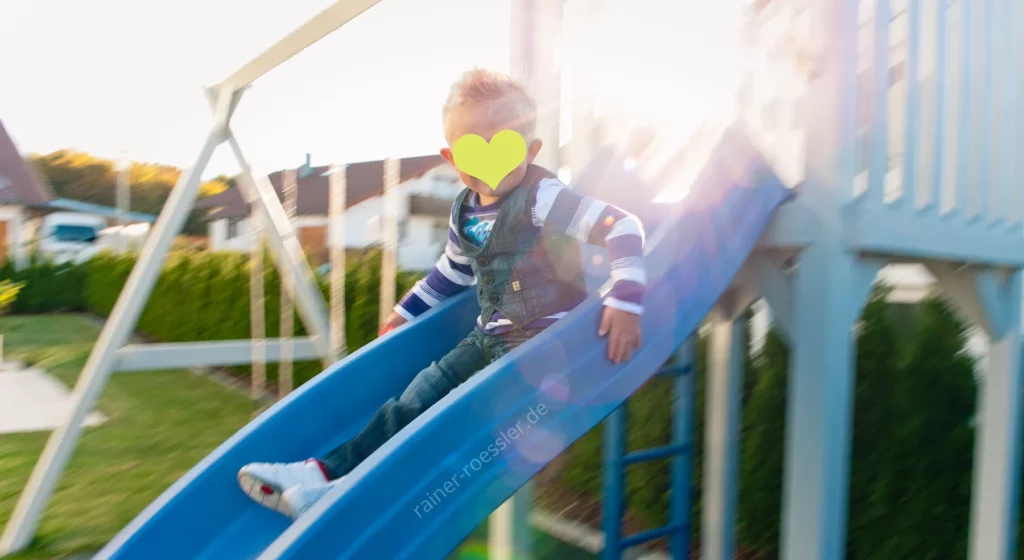 Kind rutsch eine Rutsche im heimischen Garten herunter. Das Gesicht ist per Herz unkenntlich gemacht.