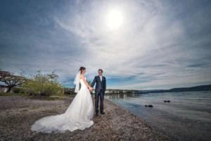 2 Menschen stehen draussen im Hochzeits-Look auf kleinen Steinen an der Promenade an einer Wasserkante. Blaue Himmel, grüne Natur und die Sonne am Himmel.