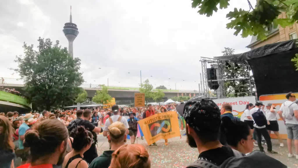 Sehr viele Menschen auf einem öffentlichen Platz draußen im Freien. Der Rheinturm hinter einem grünen Baum. Ein Gruppe läuft lachend mit einem Banner vorbei mit der Aufschrift Autistic Pride. Neurodivergent Pride. Darauf ist das unendlich Symbol in Form einer 8 zu sehen. Diese ist in Regenbogenfarben ausgemalt.