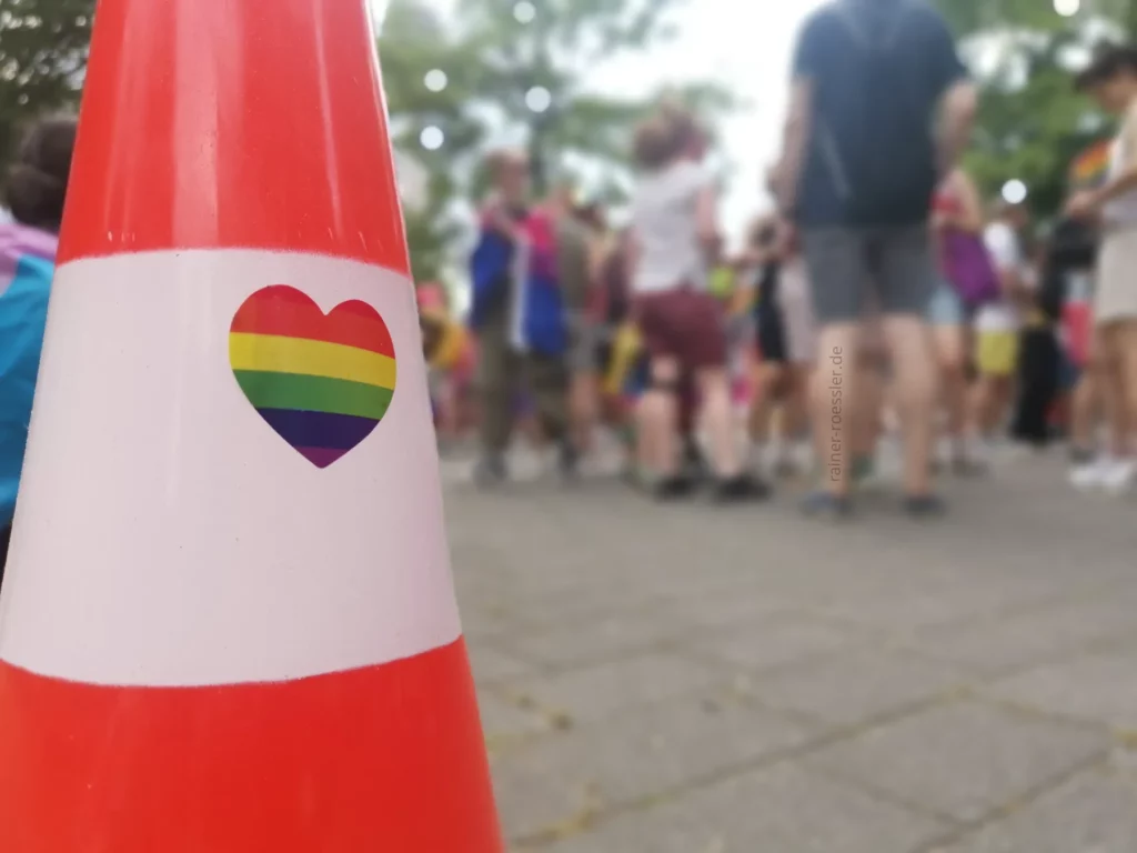 Pylone mit einem Aufkleber in Herzform und Regenbogenfarbe. Dahinter unscharf einige Menschen. Und etwas Natur.
