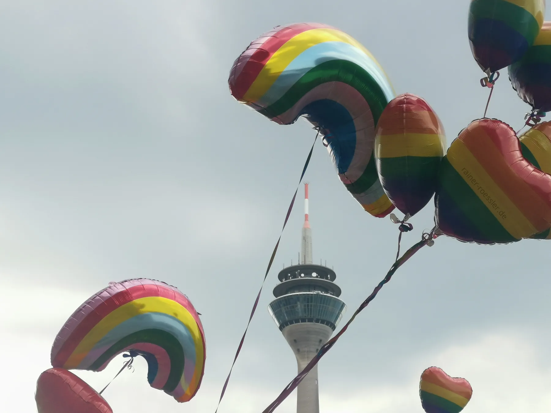 Einige Luftballone in Regenbogenfarbe. Darunter in Form von Herzen und Regenbogen. Dahinter der Rheinturm und bewölktem Himmel. Am hellen Tag.