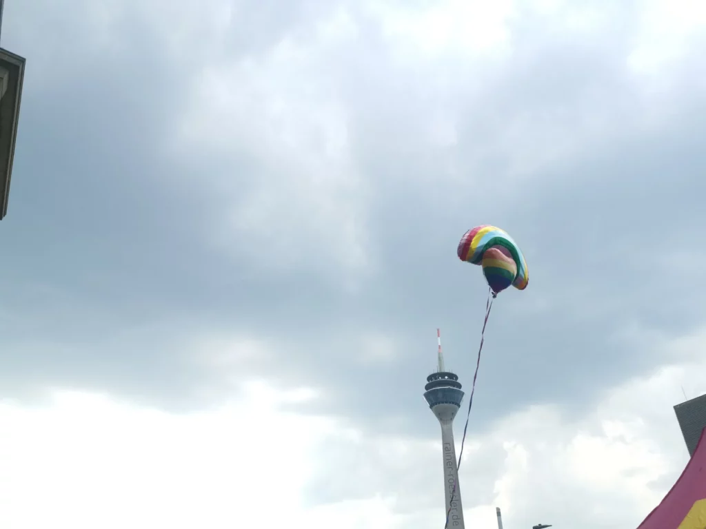 Wolken bedeckter Himmel. Rheinturm am hellen Tag. 2 Luftballone in Regenbogenfarbe in Form von einem Herz und Regenbogen davor