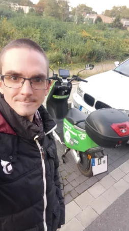 Selfie von einem Mensch stehend vor einem Moped draußen. Im Hintergrund ist grüne Natur zu sehen.