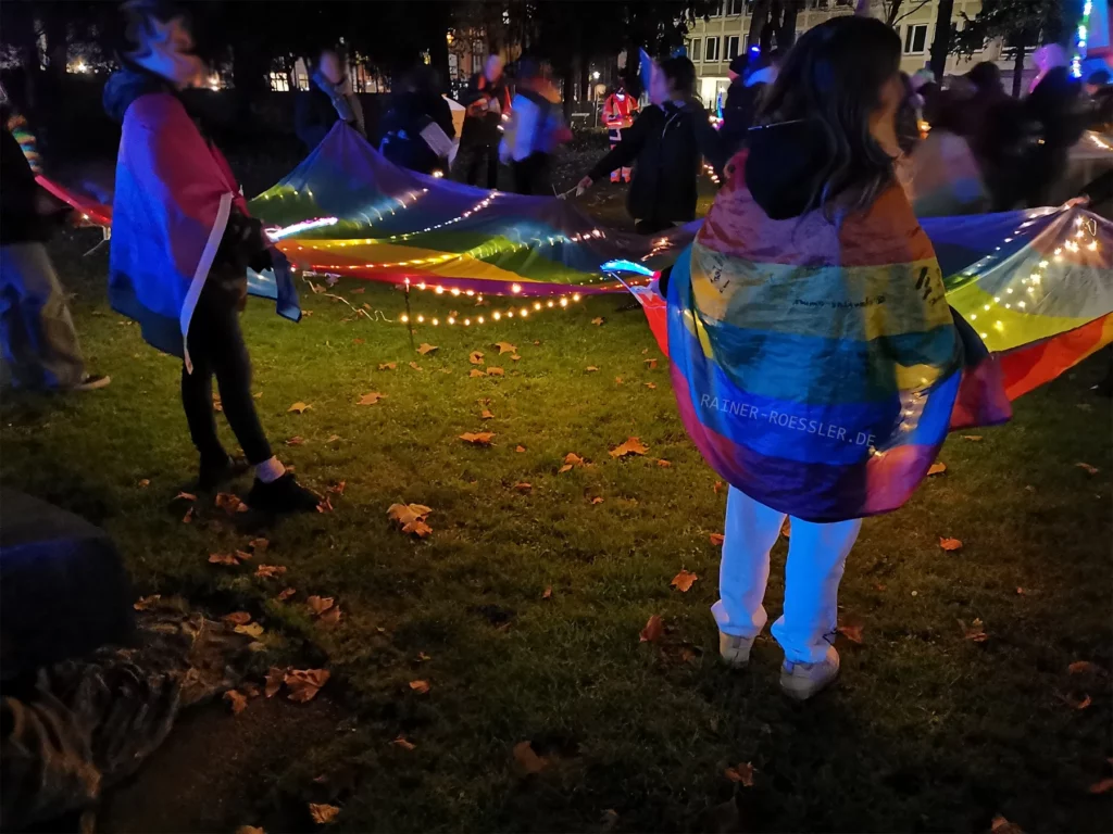 Menschen stehen im dunkeln auf einer grünen Wiese mit einer von einer Lichterkette beleuchtet Regenbogenflagge.