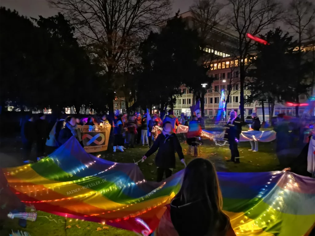 Es ist dunkel. Einige Menschen stehen draußen auf einer grünen Wiese mit bunter Ausrüstung. Eine Lichterkette ist über eine regenbogen Stoff gespannt, welche Menschen in der Luft halten. Im Hintergrund ist ein Banner zu sehen mit dem Zeichen für Neurodivergent