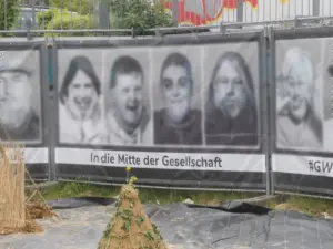 Banner an einem Gerüst. Darauf 4 Gesichter, welche digital etwas anonymisiert wurden. Darunter die Worte 