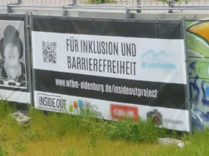 Ein Gerüst mit einem Banner steht draußen im grünen an einer Mauer aus Beton. Auf dem Banner steht 