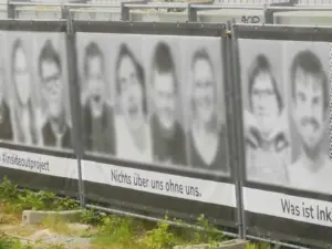 Banner auf einem Gerüst. 4 Gesichter, welche digital anonymisiert wurden. Darunter steht 