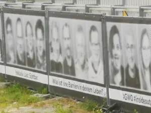 Mehrere Banner an einem Metallgerüst. Auf einem Banner in der Mitte sind 4 Gesichter, welche digital anonymisiert wurden. Darunter steht 