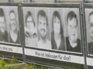 Ein Banner an einem Metallgerüst draußen an einem öffentlichen Platz. 4 Gesichter, welche digital etwas anonymisiert sind, sowie der Text darunter 
