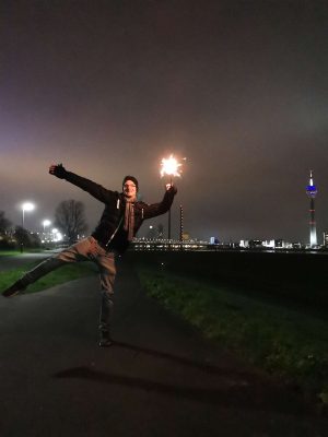 Hübscher Mann auf einem Bein balancierend am Silvesterabend mit einer Wunderkerze am Rheinufer Düsseldorf fotografiert.