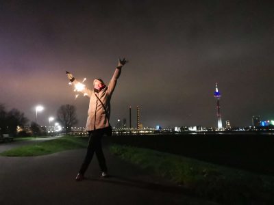 Lifestyle-Shooting - Junge Frau im grauem Mantel posiert mit Wunderkerze am Silvesterabend vor der Skyline Düsseldorf's