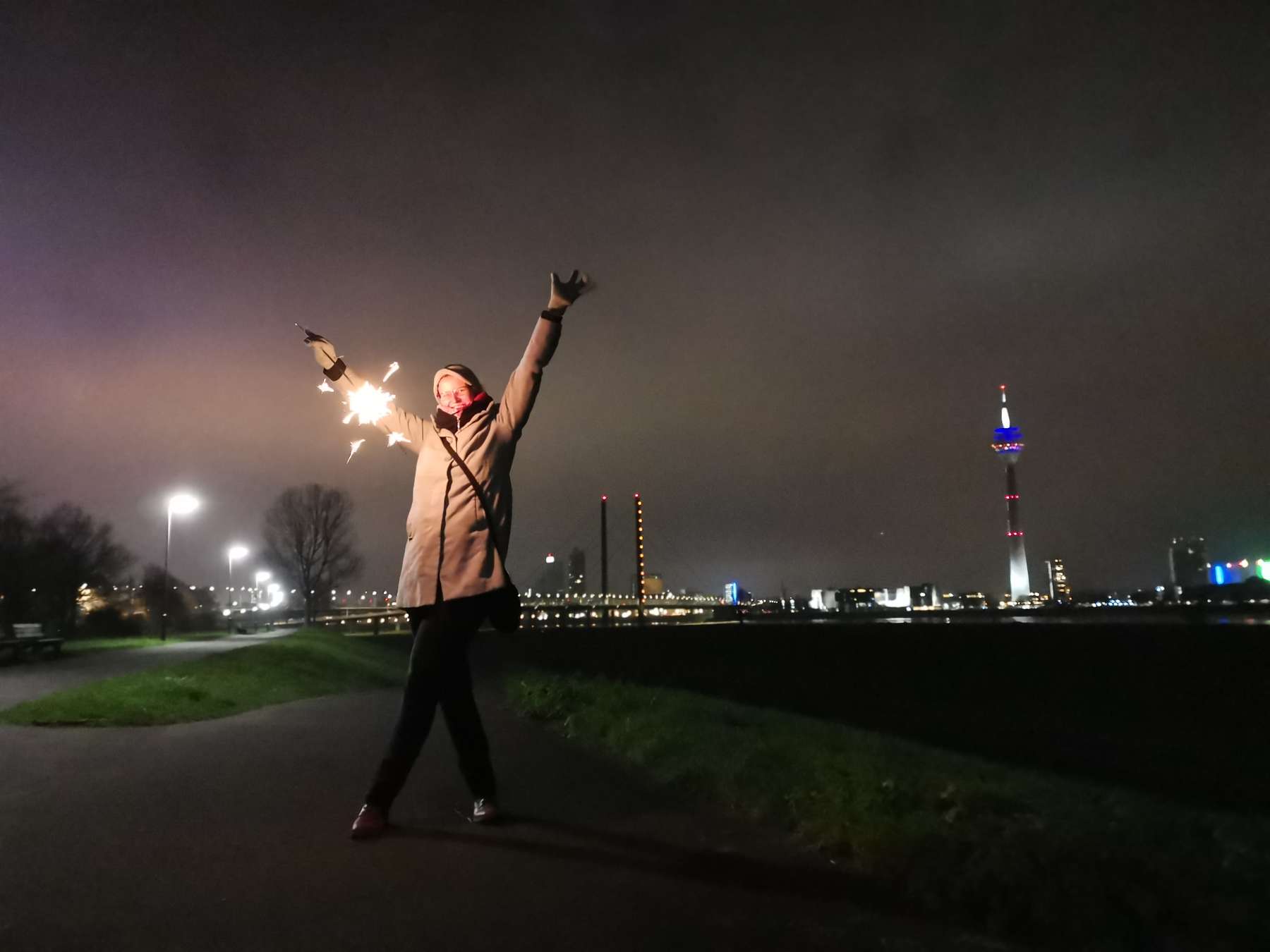 ​Lifestyle-Shooting - ​Junge Frau im grauem Mantel posiert mit Wunderkerze ​am Silvesterabend vor der Skyline ​Düsseldorf's