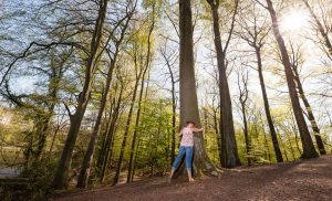 Naturverbundene junge Frau post für ein Fotoshooting in der Margarethenhöhe in inniger Umarmung mit einem Baum inmitten anderer Bäume im Stadtwald der Margarethenhöhe inmitten der Stadt Essen